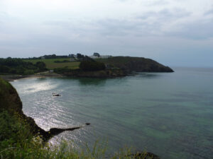 Vue depuis le GR34 sur une petite anse aux eaux vert émeraude, avec une plage en arc de cercle, des falaises rocheuses et un promontoire boisé surmonté de bâtiments, sous un ciel nuageux, presqu'île de Crozon