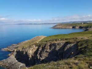 Vue panoramique depuis le GR34 sur des falaises schisteuses et une lande côtière verdoyante, avec le sentier balisé serpentant au bord des falaises et la baie de Douarnenez s'étendant à l'horizon sous un ciel bleu