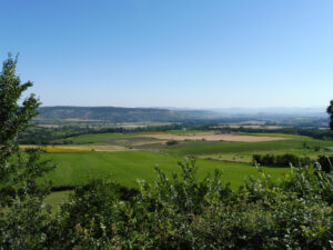 ue panoramique sur une vaste plaine agricole verdoyante en Auvergne, encadrée de végétation au premier plan, avec des collines et des sommets montagneux dans la brume à l'horizon, sous un ciel bleu dégagé.