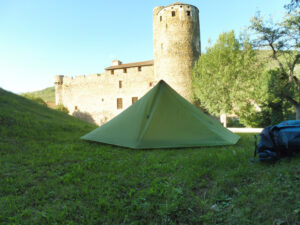 Tente de randonnée verte de type pyramidal dressée sur une pelouse, devant le château médiéval de Saint-Gervazy et sa tour ronde en pierre, en soirée d'été.