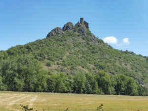 Ruines d'un château médiéval au sommet d'un piton rocheux boisé, vues depuis une prairie fauchée dans la vallée de la Couze d'Issoire, sous un ciel bleu estival.