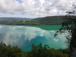 Vue en surplomb sur le lac de Chalain dans le Jura, aux eaux turquoise-émeraude, encadré de collines boisées et de cimes d'arbres en premier plan. Le ciel nuageux se reflète fidèlement sur la surface calme et lisse du lac.