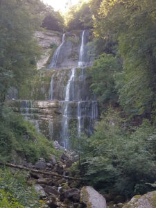 Vue frontale de la cascade de l'Éventail dans la vallée du Hérisson, Jura. L'eau dévale en plusieurs filets sur une haute paroi de calcaire étagée, encadrée d'une végétation forestière dense. Au premier plan, un lit de rochers et de troncs.