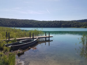 Vue sur le lac Narlay depuis sa rive, aux eaux turquoise transparentes. Au premier plan, une vieille barque en bois sombre est amarrée contre un petit ponton en bois, entouré de roseaux. En arrière-plan, une colline entièrement couverte de forêt se reflète dans l'eau sous un ciel bleu d'été.