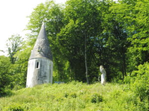 Ancienne tour ronde en pierre à toit conique, partiellement envahie par la végétation, accompagnée d'une grande sculpture en bois représentant une chouette, au milieu d'un sous-bois verdoyant en Pays de Caux, Normandie.