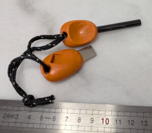 allume-feu à friction de type fire starter, à poignée ovale orange et cordelette noire, posés sur une surface blanche devant une règle graduée : l'un présente la tige en ferrocérium noire, l'autre la racle métallique argentée intégrée dans la poignée.