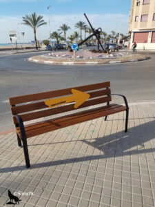 Banc en bois avec une flèche jaune « Camino de Santiago », devant un rond-point orné d'une grande ancre marine et bordé de palmiers, au bord de mer à Sagunto