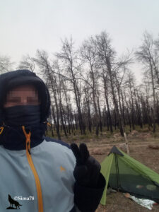 Selfie d'un pèlerin emmitouflé dans une veste à capuche, cagoule et gants noirs, faisant le signe V de la victoire devant sa petite tente verte ultralight plantée au milieu d'une forêt de pins calcinés par un incendie, par temps gris et venteux