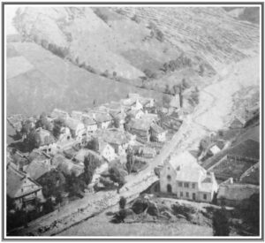 Vue aérienne en noir et blanc d'un village de montagne groupé dans un vallon, entouré de prairies et de versants, vers 1880.