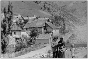 Photographie ancienne en noir et blanc : deux adultes et un enfant debout au bord d'un chemin de village alpin, avec des maisons et un versant montagneux en arrière-plan.