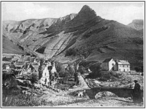 Photographie ancienne en noir et blanc montrant les ruines du village de Chaudun après son abandon : maisons démolies ou éventrées à gauche, église et maison curiale encore debout à droite, pont de pierre au premier plan, massif montagnard en arrière-plan.