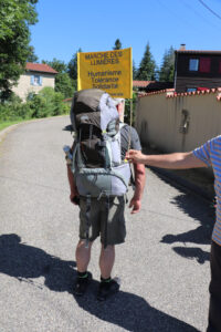 Hubert Constancias de dos, portant un grand sac à dos de randonnée, sur une route de village. Une bannière jaune arbore les mots « Marche des Lumières – Humanisme, Tolérance, Solidarité – Août 2024 ».