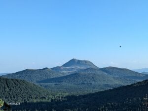 Vue panoramique sur le Puy de Dôme et ses volcans voisins couverts de forêts, avec une montgolfière dans le ciel bleu.