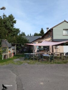 Terrasse d'un bar-restaurant de village en Auvergne, avec tables et chaises sous une voile d'ombrage rose, barbecue extérieur et enseigne "Bar" visible sur la façade.