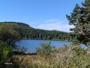 Lac de maar volcanique aux eaux bleu profond, encadré de forêts de conifères et d'une végétation rivulaire, avec un cône volcanique visible à l'horizon sous un ciel bleu.