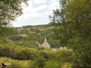 Vue depuis le sentier sur la basilique romane Notre-Dame d'Orcival, édifice en pierre volcanique grise émergeant d'un écrin de forêts verdoyantes au fond d'une vallée auvergnate.