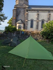 Deux tentes de bivouac légères installées sur la pelouse au pied de l'église romane en pierre de Volvic d'Olby, tente pyramidale verte au premier plan et tente dôme kaki derrière.