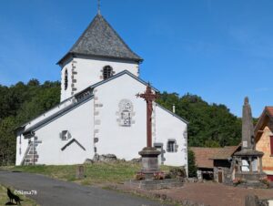 Église de village auvergnate à façade blanche et encadrements en pierre volcanique, avec une haute croix de mission en fer forgé et un monument aux morts sur la place, sous un ciel bleu vif.