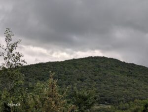 Flanc d'un cône volcanique entièrement boisé se découpant sous un ciel dramatique chargé de nuages gris anthracite, avec une percée lumineuse blanche à la ligne de crête.