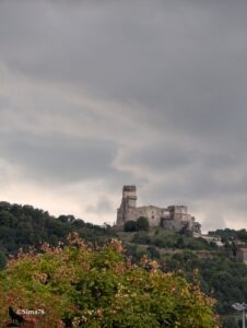 Ruines d'un château médiéval perché sur une colline boisée, tour crénelée et murailles en partie effondrées se détachant sur un ciel d'orage dramatique, feuillages aux premières teintes automnales au premier plan.