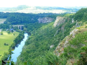 Vue plongeante depuis un belvédère rocheux sur la vallée encaissée de l'Orne, avec les falaises de grès armoricain à droite, le viaduc ferroviaire de Clécy en arrière-plan et la campagne normande s'étendant à l'horizon, en Suisse Normande.