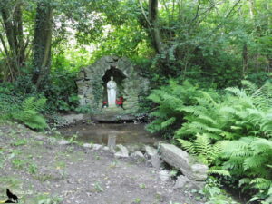 Petite grotte votive en pierres sèches abritant une statue de saint tenant l'Enfant Jésus, ornée de fleurs rouges, entourée de fougères luxuriantes et de végétation dense en sous-bois, en Suisse Normande, Normandie.