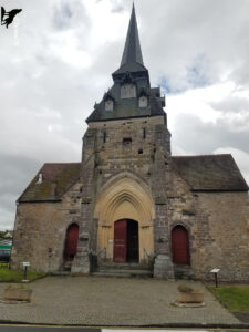Façade frontale de l'église Saint-Laurent de Clécy, édifice roman en pierre de granit avec un portail ogival en pierre de taille claire, surmonté d'un clocher à flèche d'ardoise, sous un ciel nuageux en Suisse Normande.