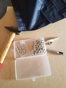 Vue de dessus sur une surface en bois, avec un kit d'œillets métalliques argentés en boîte plastique transparente à deux compartiments, accompagné d'un poinçon et d'un poseur d'œillets en métal ciselé, un marteau à manche en bois, et un poncho en nylon bleu marine en arrière-plan.