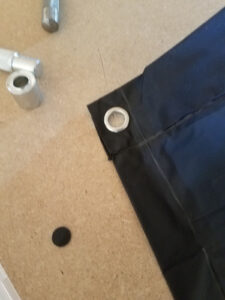 Gros plan sur un coin de poncho en nylon bleu marine avec un œillet métallique argenté fraîchement posé sur le bord renforcé, avec le poinçon et le poseur d'œillets en métal posés à gauche sur la surface en bois, et le disque de tissu découpé visible au sol.