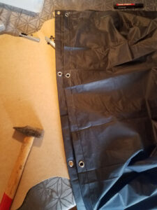 Vue d'ensemble du poncho en nylon noir transformé en tarp, avec une rangée de cinq œillets métalliques argentés régulièrement espacés le long du bord renforcé cousu, posé sur une planche en bois, avec le marteau, les outils de pose et un marqueur rouge visibles à côté.