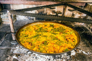 Grande poêle à paella métallique contenant une paella valenciana au riz safran avec haricots verts et légumes, posée sur des braises dans un foyer rustique en briques et fer forgé.