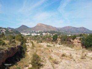 Paysage aride de garrigue méditerranéenne avec un lit de rivière asséché au premier plan, un village blanc niché au pied de collines boisées au milieu, et une mesa rocheuse aux teintes ocre dominant l'horizon sous un ciel bleu