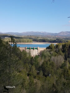 Vue plongeante sur un barrage-poids en béton aux vannes métalliques vertes, retenant un lac de retenue aux eaux bleues partiellement à nu, encadré de pinèdes et de collines, avec des éoliennes visibles sur la crête en arrière-plan