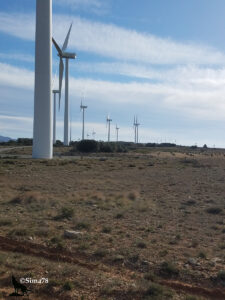 Alignement de nombreuses éoliennes blanches sur un plateau aride aux herbes rases et aux terres rougeâtres, sous un ciel bleu strié de cirrus, avec un mât d'éolienne au premier plan en très gros plan