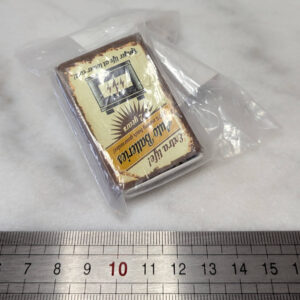 Petite boîte d'allumettes à décor rétro style "Auto Batteries", glissée dans un sachet plastique transparent zippé et posée sur une surface blanche devant une règle graduée, mesurant environ 5 cm de long.