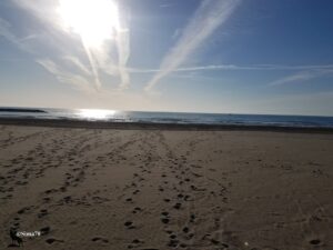 Plage de sable déserte à contre-jour, parsemée d'empreintes de pas, face à une mer Méditerranée scintillante sous un soleil matinal et un ciel bleu strié de traînées nuageuses