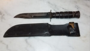 Poignard militaire américain de marque Camillus posé sur fond de marbre blanc, lame à fuller central patinée et noircie, garde en croix, poignée en rondelles de cuir empilées, présenté avec son fourreau en cuir noir à système de rétention à bouton-pression, montrant des signes d'usure et de patine d'époque.