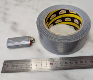 Rouleau de ruban adhésif toilé gris Pattex d'environ 10 cm de diamètre posé à côté d'un briquet BIC miniature entièrement recouvert de plusieurs tours du même adhésif gris, sur une surface blanche avec une règle graduée en centimètres.