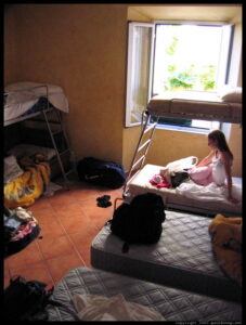 Dortoir d'auberge de jeunesse avec trois lits superposés en métal, un matelas au sol, carrelage en terre cuite, sacs à dos et équipements de randonnée dispersés, fenêtre ouverte sur végétation extérieure.