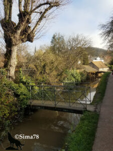 Passerelle métallique enjambant un petit cours d'eau à Chevreuse, bordée d'arbres dénudés en hiver, avec des maisons de village et un clocher d'église visibles en arrière-plan. Photo ©Sima78.