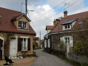 Ruelle étroite traversant un hameau de la Vallée de Chevreuse, bordée de maisons en pierre meulière aux volets bruns et blancs, dont l'une est recouverte de végétation grimpante. Ciel nuageux de fin d'hiver. Photo ©Sima78.