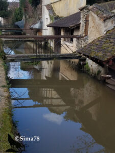 Vieilles maisons en pierre aux facades patinées se reflétant dans les eaux calmes de l'Yvette, avec une passerelle métallique rouillée enjambant le cours d'eau au cœur de Chevreuse. Photo ©Sima78.