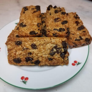 Cinq barres de céréales maison aux flocons d'avoine et raisins secs, dorées et généreuses, présentées sur une assiette blanche à motifs de cerises rouges, posée sur un plan de travail en marbre.