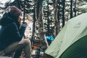 Randonneuse assise de profil près d'une tente MSR verte en forêt de conifères, vêtue d'une veste bleue et d'un bonnet mauve, tenant une tasse fumante entre ses mains gantées, avec un réchaud de randonnée au sol et un sac à dos bleu en arrière-plan.