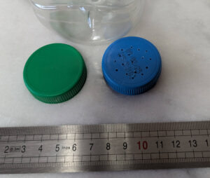 Gros plan sur deux bouchons à vis en plastique d'environ 4 cm de diamètre posés côte à côte devant une règle graduée, au pied d'une bouteille PET transparente : un bouchon vert lisse et hermétique, un bouchon bleu percé d'une quinzaine de petits trous irréguliers sur toute sa surface.