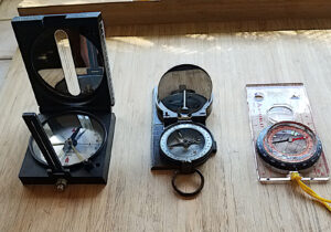 Trois boussoles posées sur une surface en bois : une boussole à miroir H. Morin-Secretan (à gauche), une boussole à prisme de type militaire (au centre), et une boussole à semelle transparente de marque McKinley (à droite).