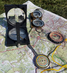 Trois boussoles de types différents et un curvimètre à molette posés sur une carte topographique IGN dépliée à l'extérieur, sur l'herbe.