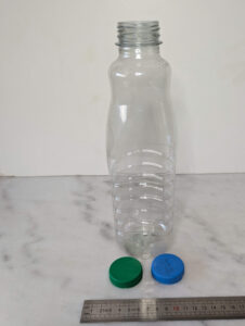 Bouteille en plastique PET transparent d'environ 75 cl à paroi striée, posée debout sur une surface en marbre devant une règle graduée (environ 28 cm de haut), avec deux bouchons à vis posés devant elle : un vert hermétique et un bleu perforé de petits trous.