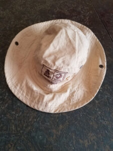 Chapeau de randonnée en coton beige posé à plat sur une surface en pierre grise, orné d'un patch brodé « Dakar » et « Partenaire Officiel », avec des œillets métalliques pour remonter les bords.