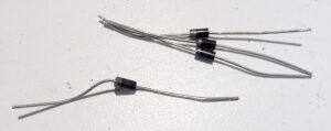 Quatre diodes électroniques à boîtier cylindrique noir DO-41 avec fils de connexion métalliques, posées en vrac sur un fond blanc, dont trois regroupées et une séparée.